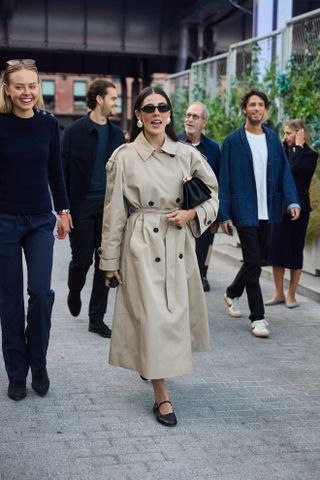 a guest attends new york spring/summer 2025 fashion week wearing a trench coat black sunglasses and black mary jane ballet flats