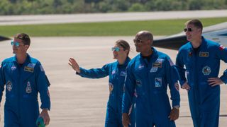 four people in blue flight suits and sunglasses smile and wave on a concrete tarmac