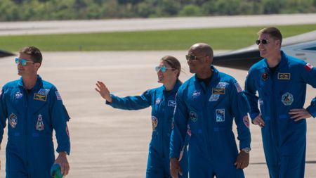 four people in blue flight suits and sunglasses smile and wave on a concrete tarmac