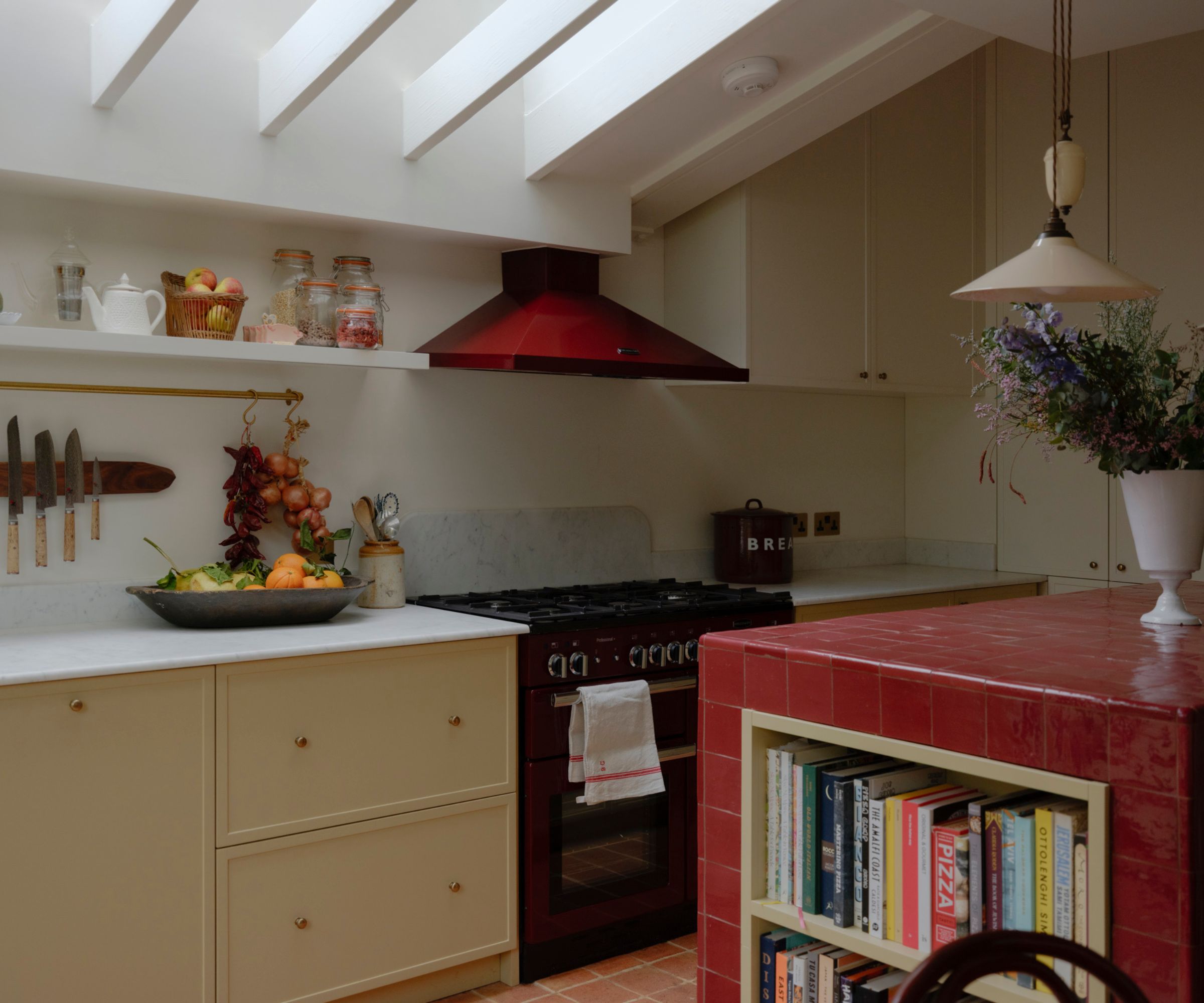 Warm cream kitchen with a red range cooker and matching red extractor hood beneath white ceiling beams. A red tiled island with built-in bookshelves stands in the foreground, while open shelves display jars, fruit and hanging onions for a homely feel.