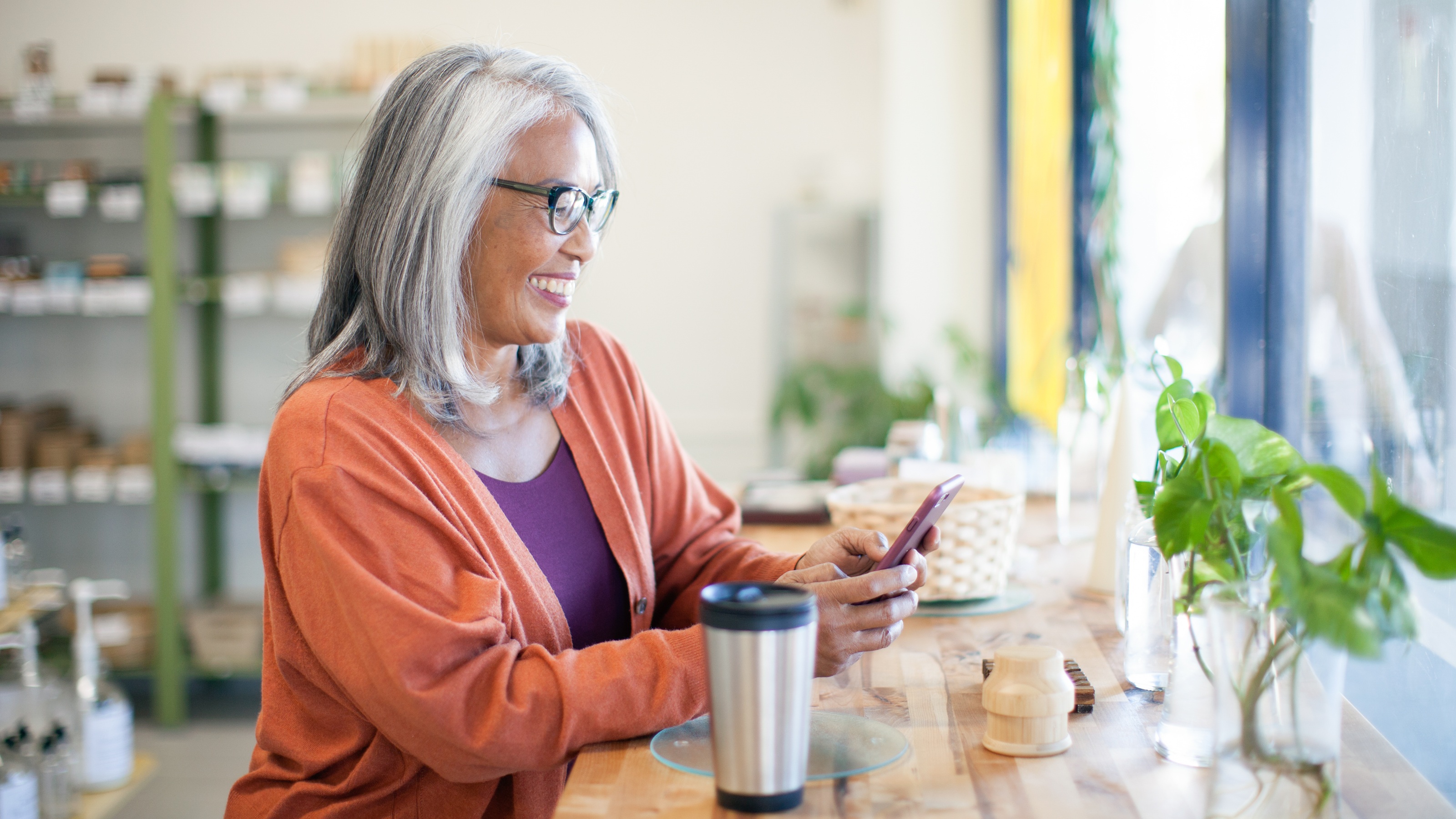 An older woman smiles while she sits in a shop and looks at her phone.