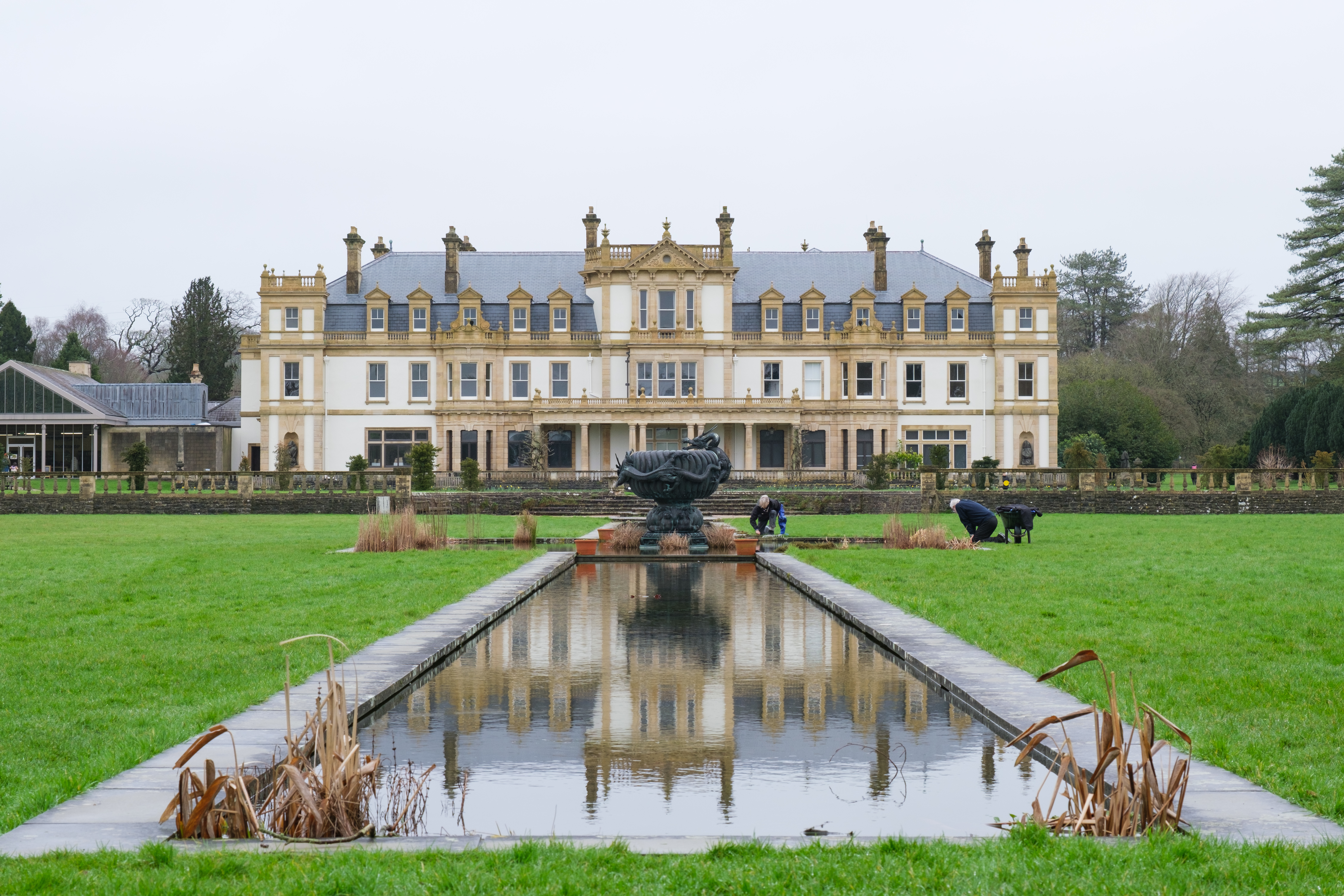 A photo of a British stately home on an overcast day, reflected in a water feature with a large sculpture in the mid-ground.