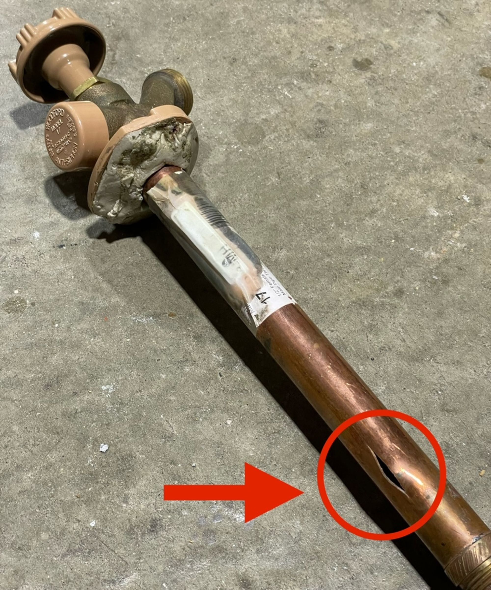 A copper pipe with a vertical gash lays on a cement floor. The photo has identified the gash with a red arrow and circle graphic. The spigot above is intact