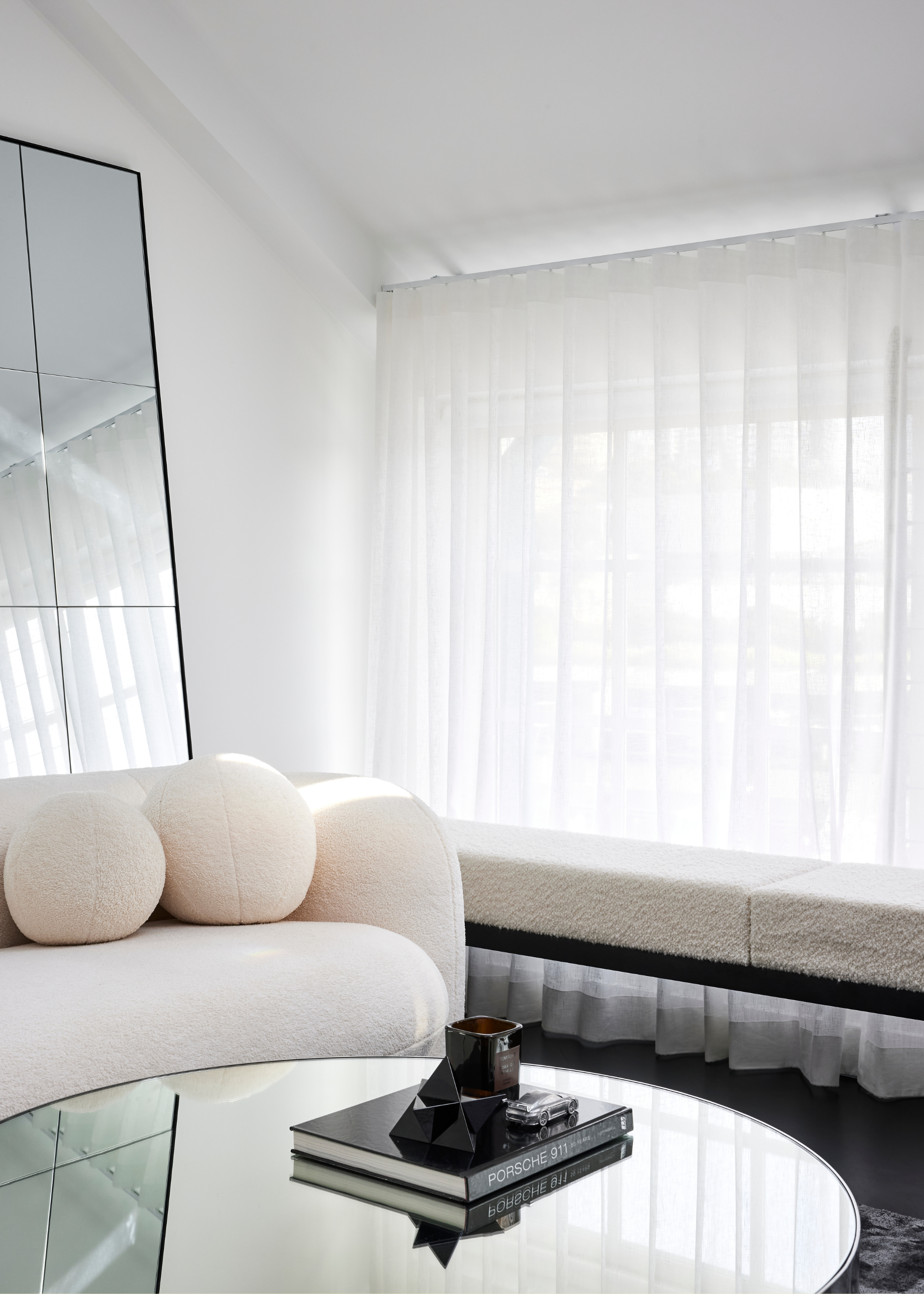 A white living room with a boucle couch, round pillows, a mirrored coffee table with a book and a scented candle