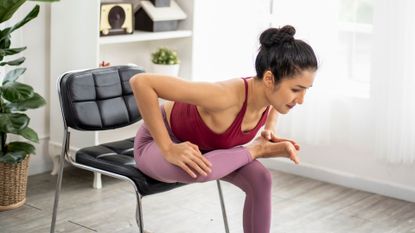 Woman demonstrating supported pigeon pose at home on chair in living room as part of a chair yoga workout
