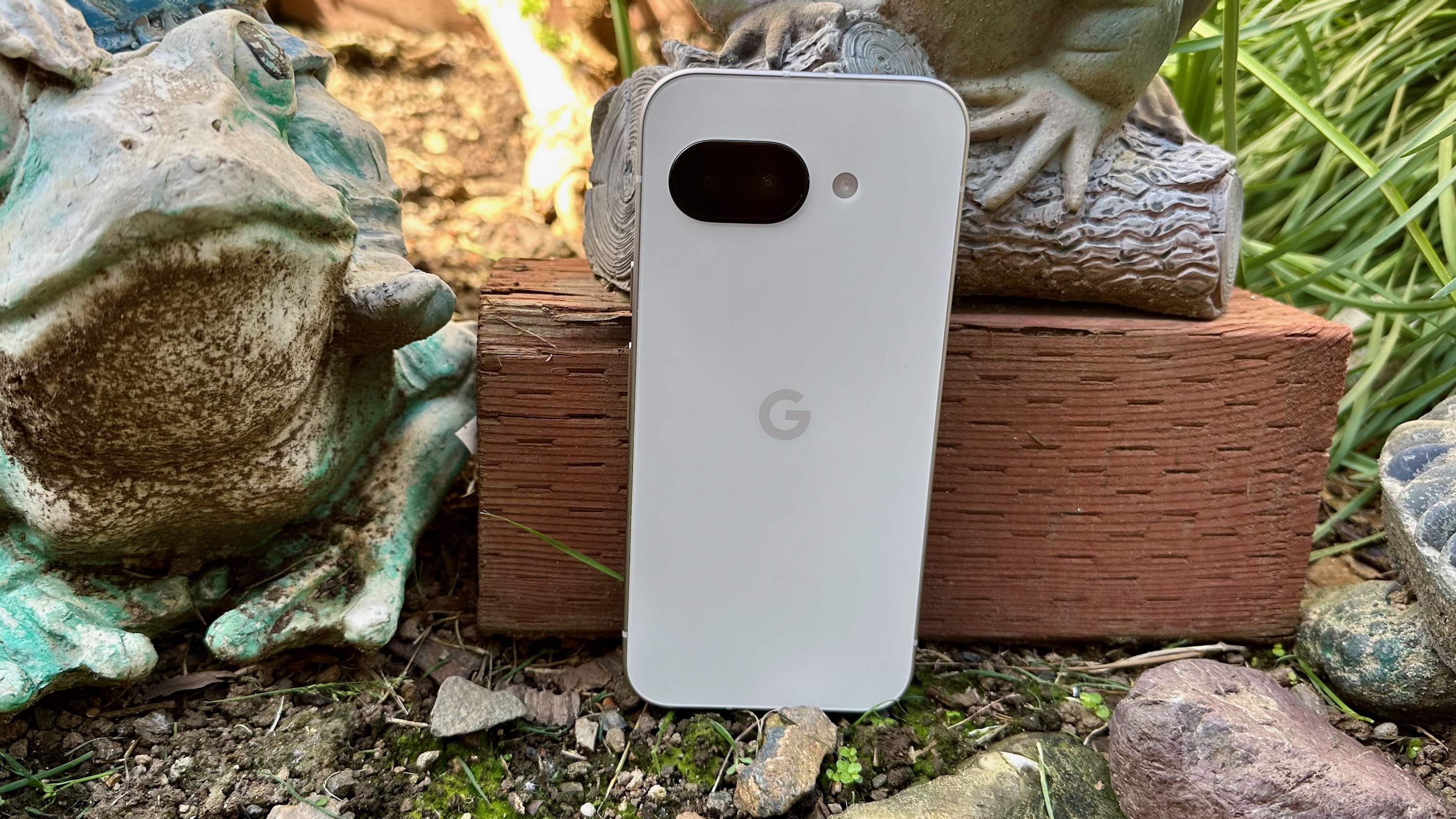 The Google Pixel 9a sitting in a garden next to a frog statue.
