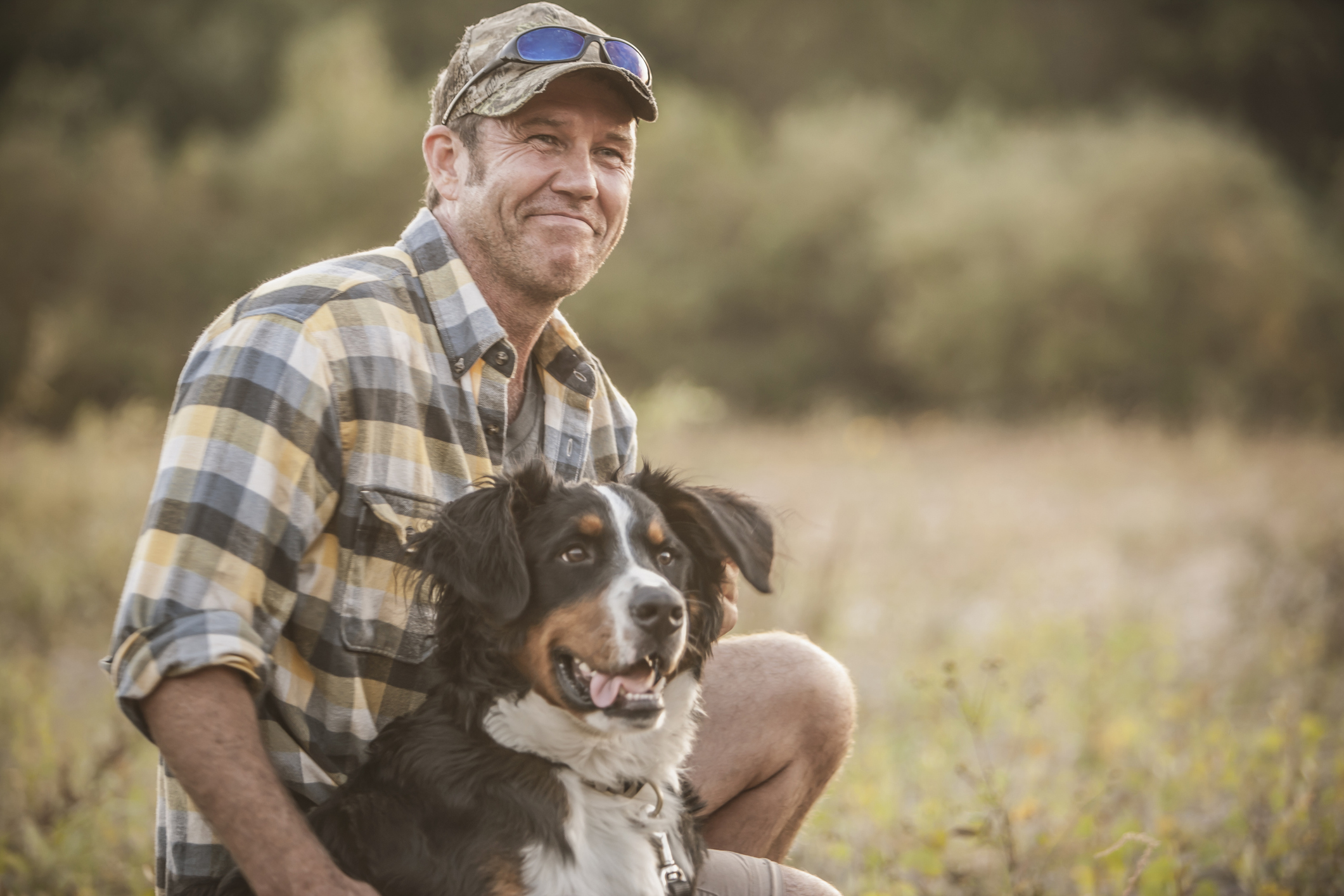 A man in his 50s kneels by his dog in a rural field. He is smiling, while his dog looks to the side.