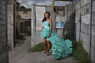 A young girl poses for the camera wearing a bright turquoise, frilly dress, framed by breeze block walls
