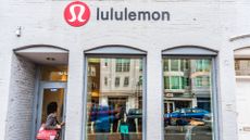 A lululemon storefront with two windows. You can see the street behind the photographer reflected in the window. A woman with a red tote bag under her arm is entering the store