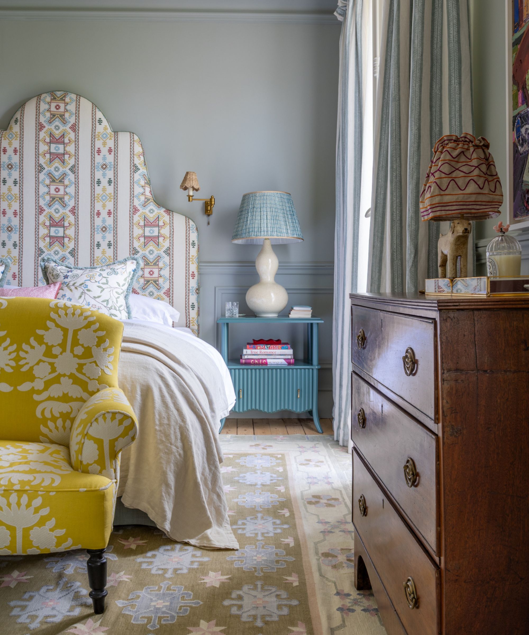 A pale blue bedroom with a tall patterned headboard, yellow armchair at the end of the bed, blue side table with a white lamp, and a muted patterned rug on wooden floors.