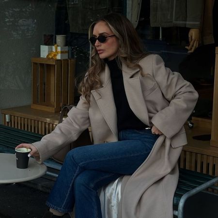 Woman wears black turtleneck jumper, beige coat and blue jeans