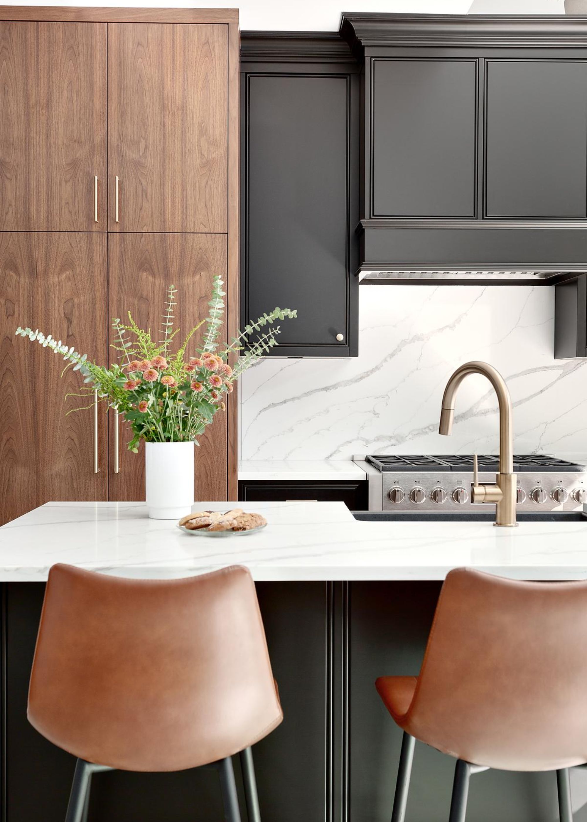 A kitchen with contrasting black and dark wood cabinets as well as a marble backsplash and a view of a stovetop. There is also a kitchen island in the middle with stools beside it as well as a vase and flowers inside.