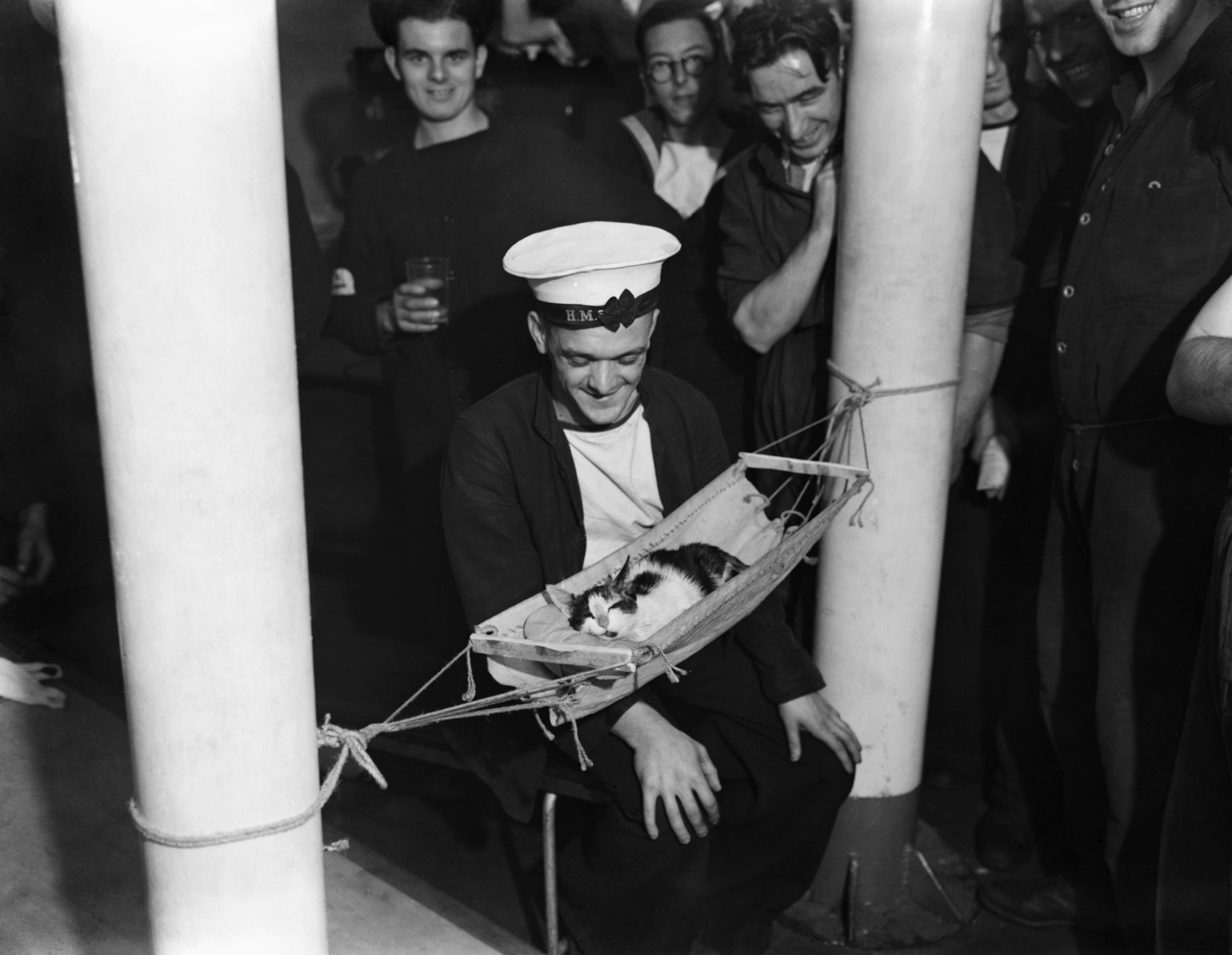 HMS Hermione, Gibraltar, 26 November 1941, A group of sailors surround the ship's cat 'Convoy', as he sleeps in a miniature hammock on board HMS HERMIONE. According to the original caption, 'Convoy' is in the ship's book and provided with full kit. The cat obtained his name because of the number of times he accompanied the ship on duty patrols, 26 November 1941. (Photo by Lt. S J Beadell/ Imperial War Museums via Getty Images)