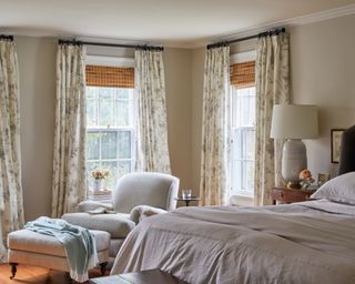 neutral bedroom with floral drapes, neutral armchair and footstool and bed with dark gray headboard
