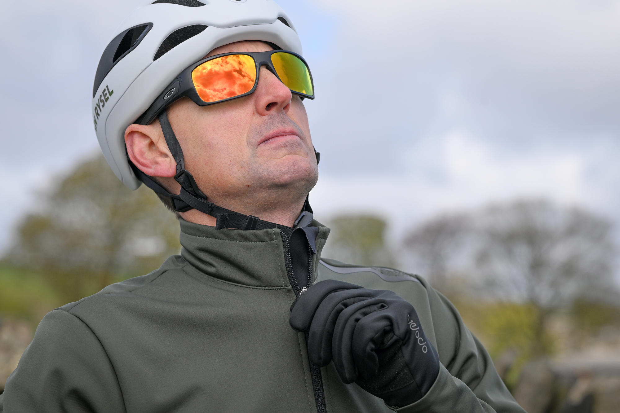 man zipping up a dark green cycling jacket wearing a white helmet, sunglasses and black gloves