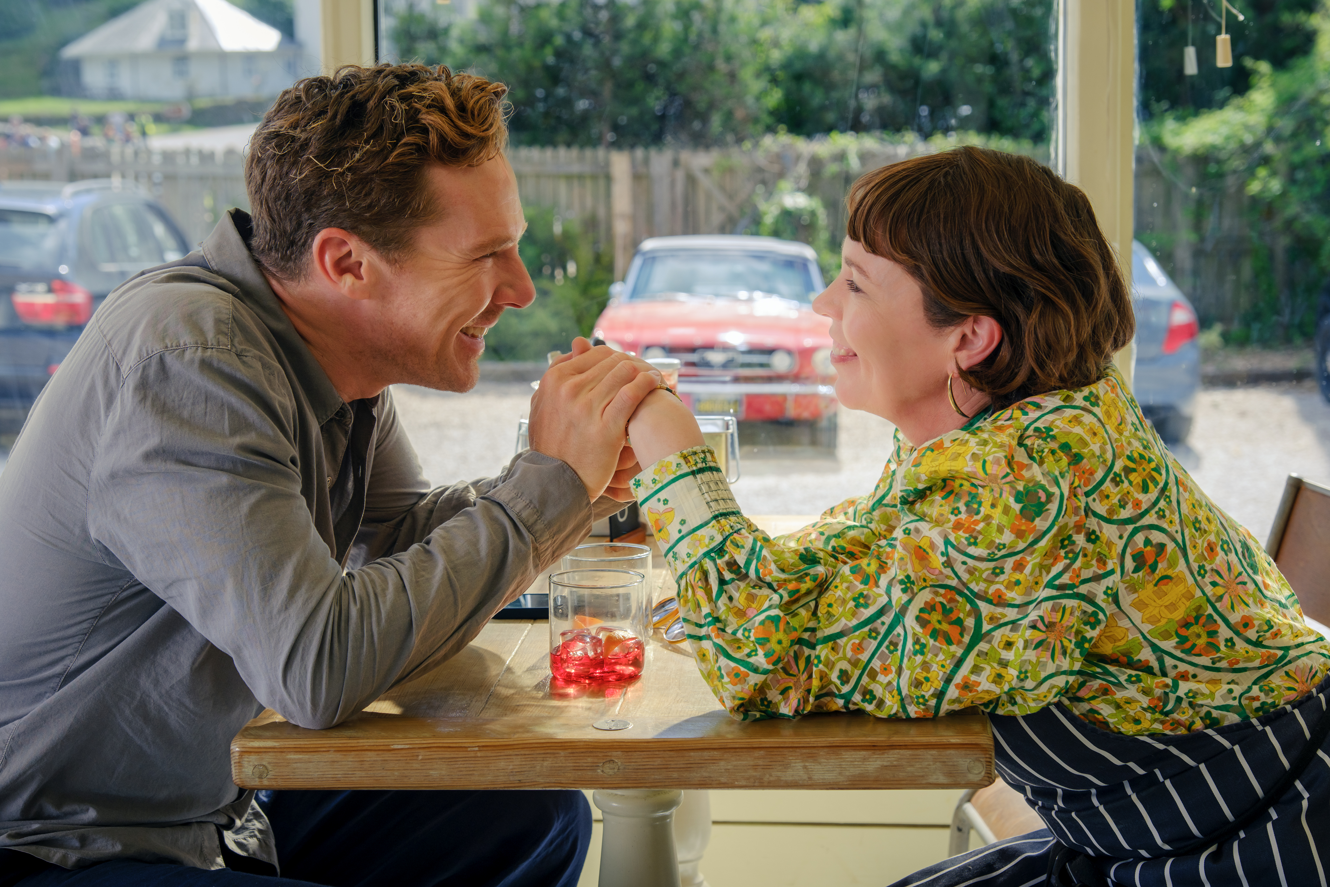 benedict cumberbatch and olivia colman hold hands over a table smiling at each other in a still from the roses