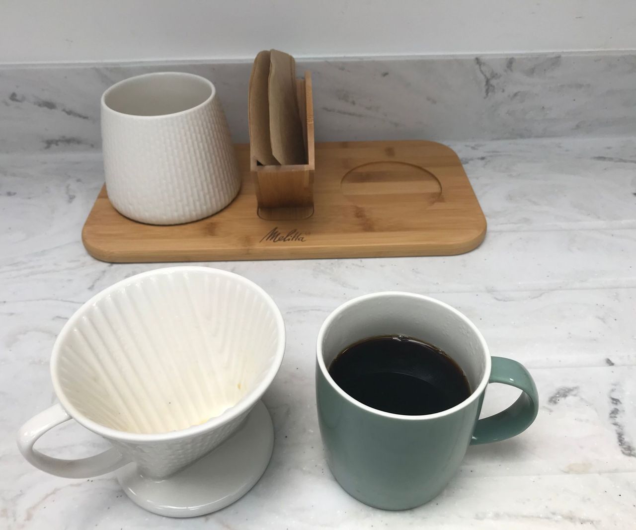 Melitta pour-over coffee maker next to a mug of black coffee on a marble kitchen counter. A wooden stand with a compartment for filters is in the background.