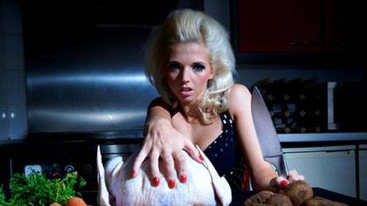 blond woman at table in dark kitchen reaching for raw chicken