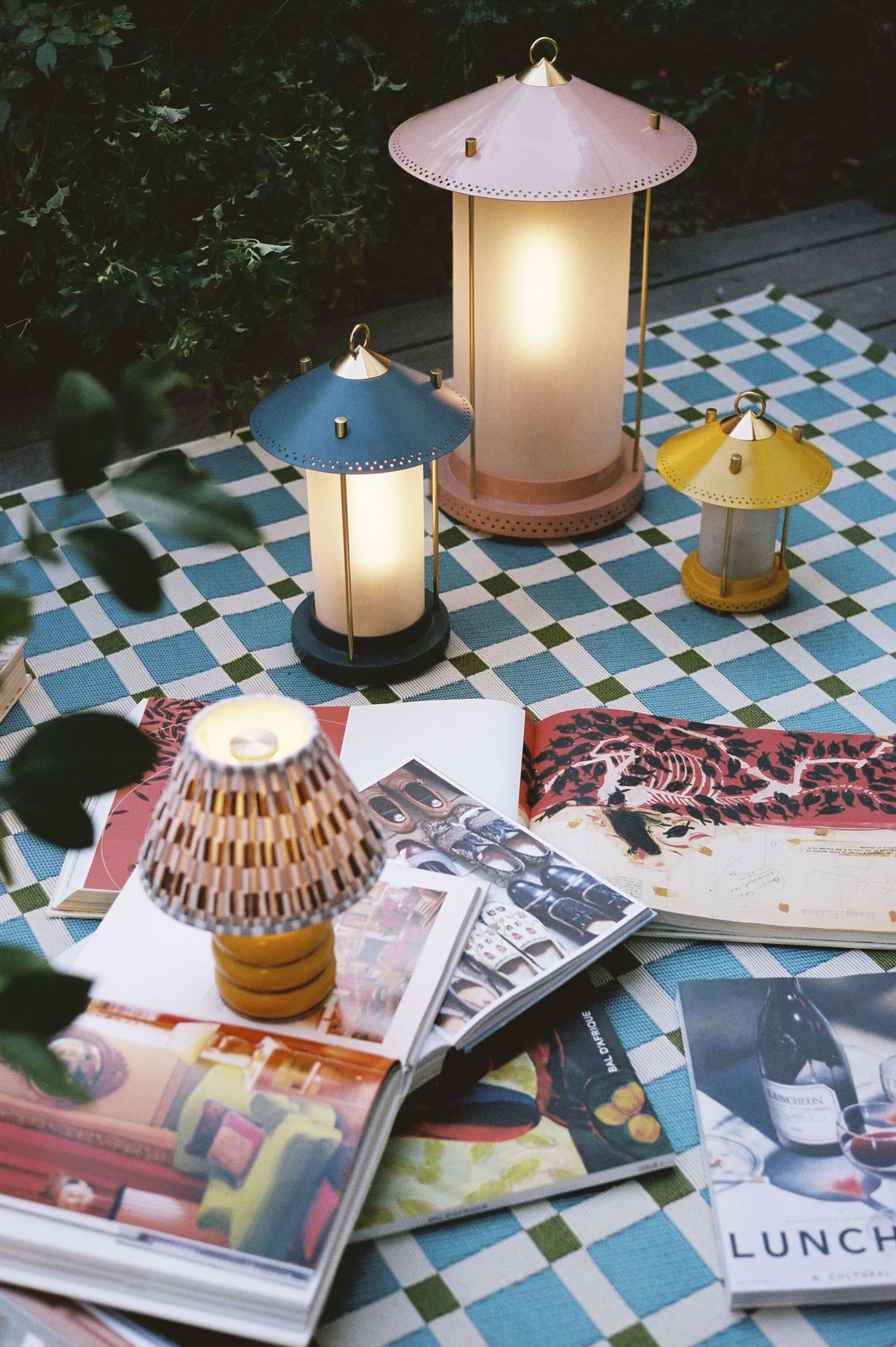 Image of three colorful lanterns outside on a blue, white, and green checkerboard blanket. There are multiple open magazines also on the blanket.