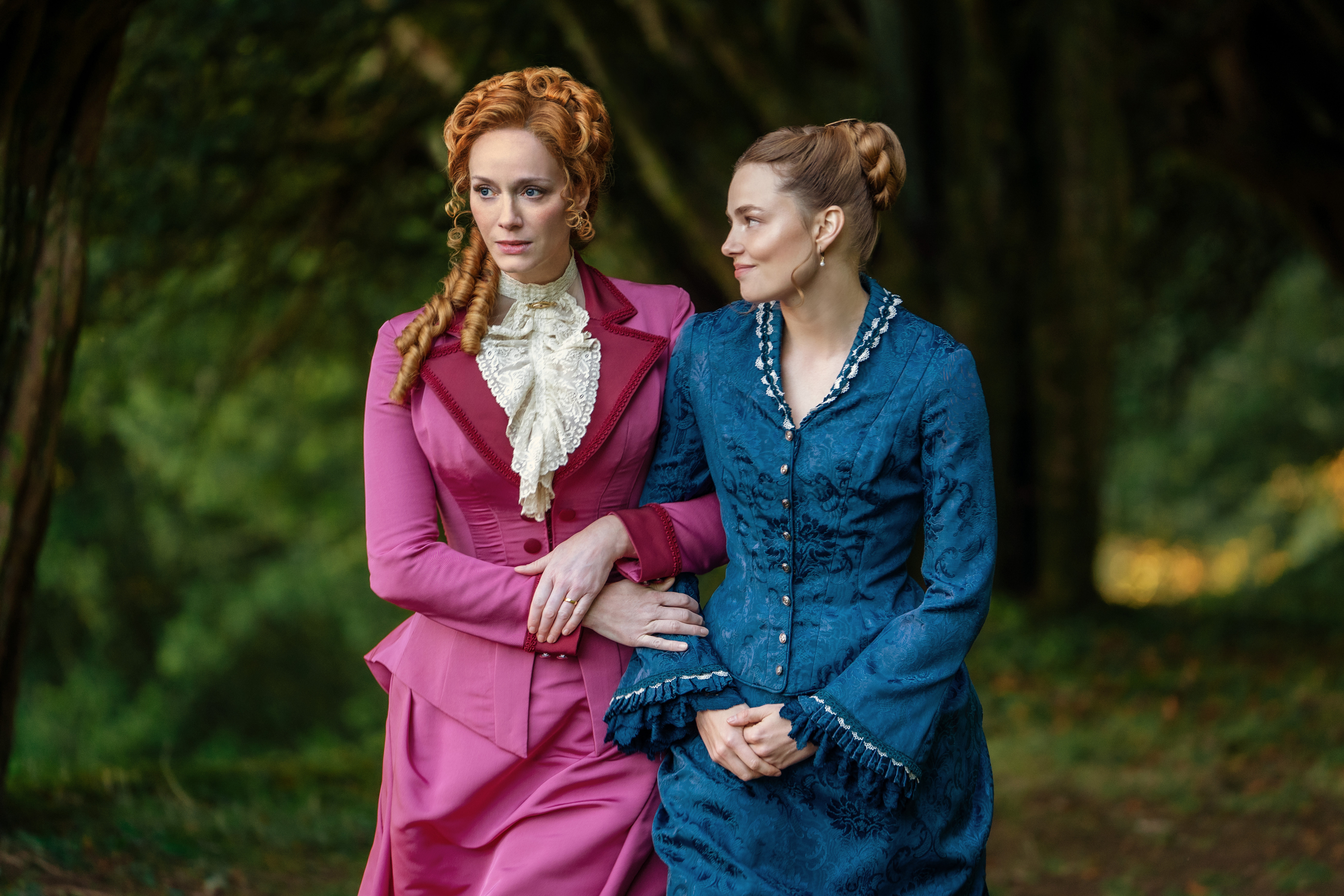 christina hendricks in a pink dress and kristine froseth in a blue dress walking through a field in the buccaneers season 2