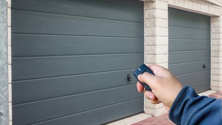 A hand holding a garage remote aimed at a double-bay garage with two distinct grey doors. 