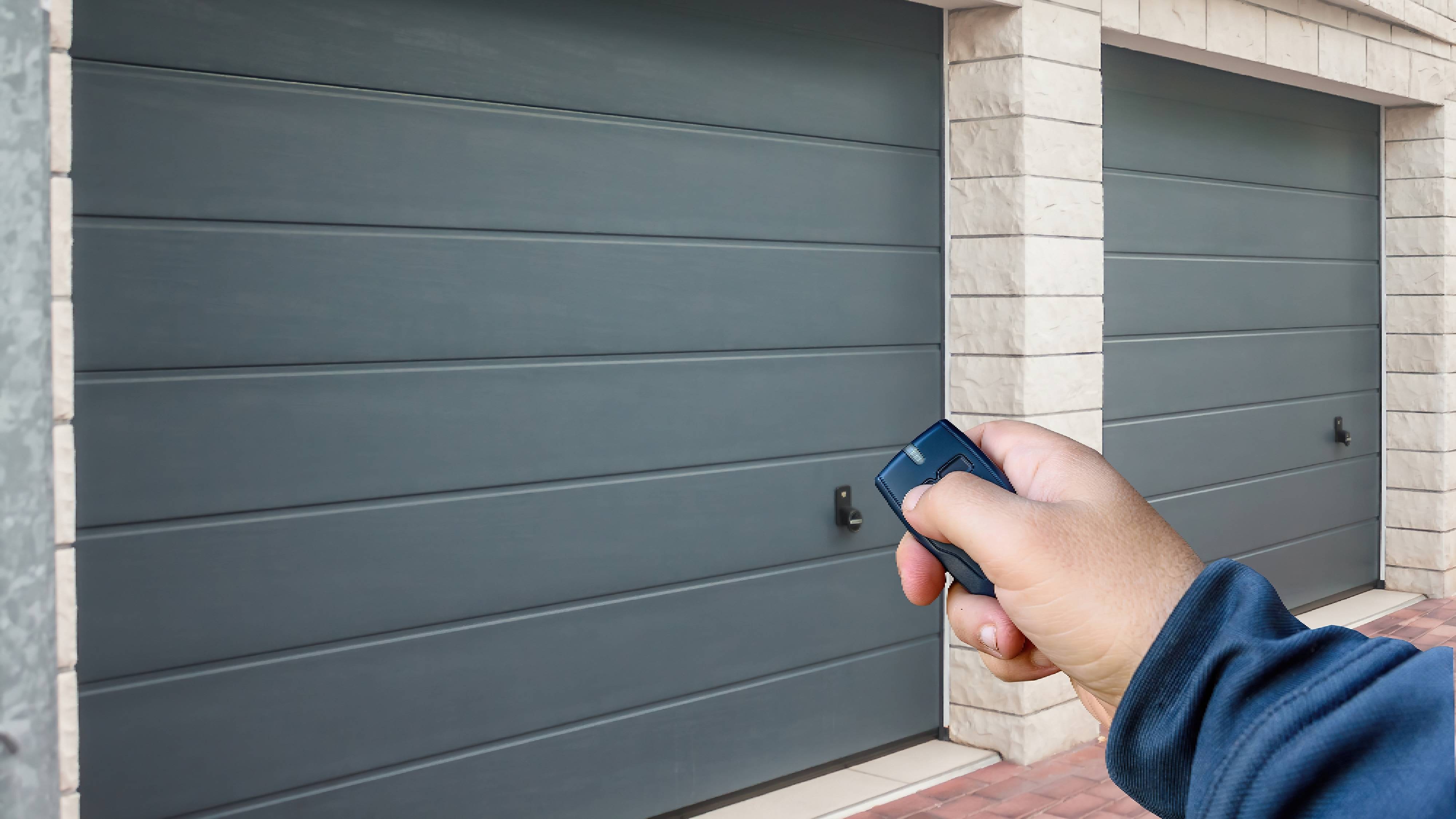 A hand holding a garage remote aimed at a double-bay garage with two distinct grey doors. 