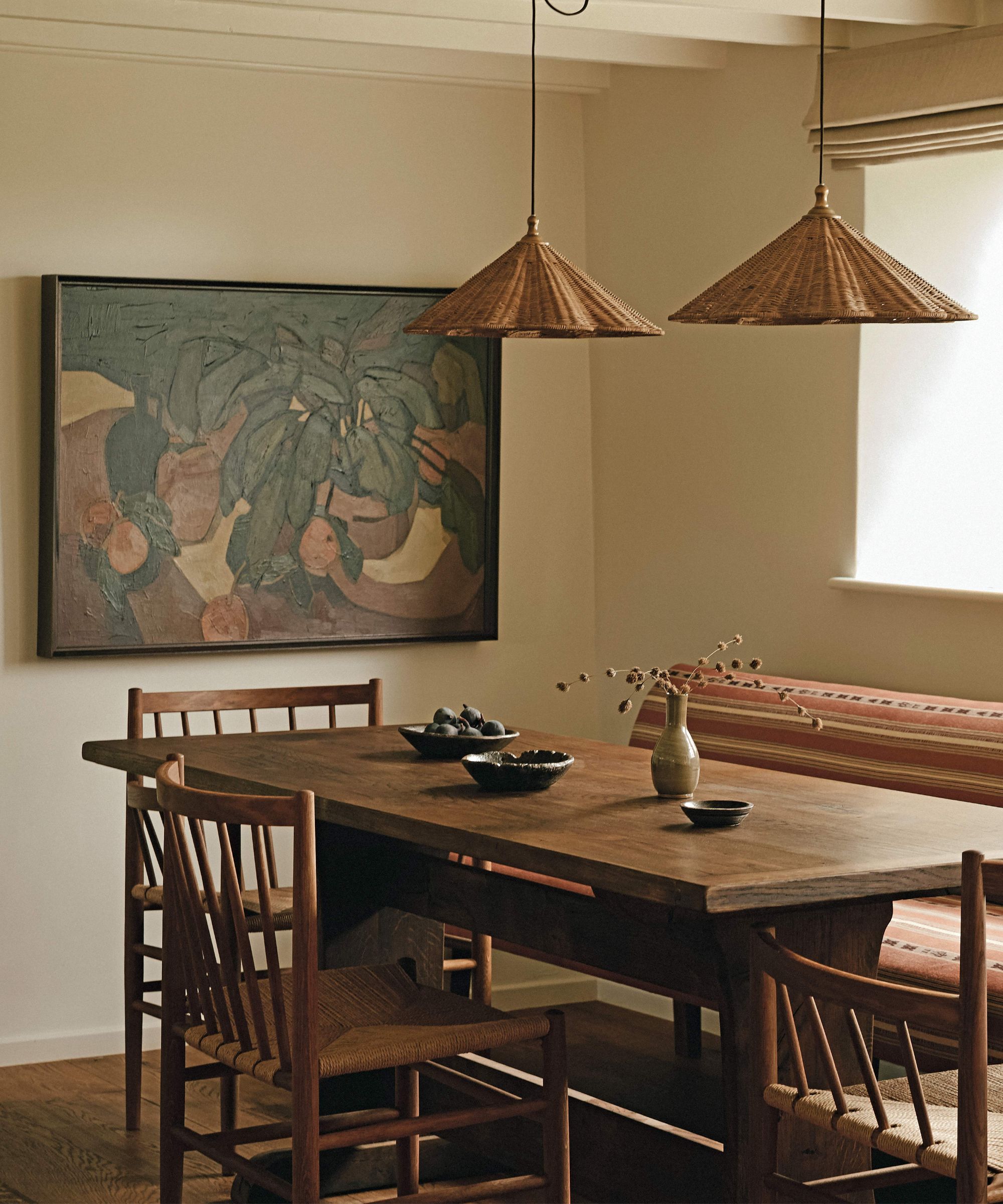 a warm neutral dining room with panelled ceiling, two rattan scalloped pendants, a banquette seat and dark wooden vintage furniture with a large painting on the wall