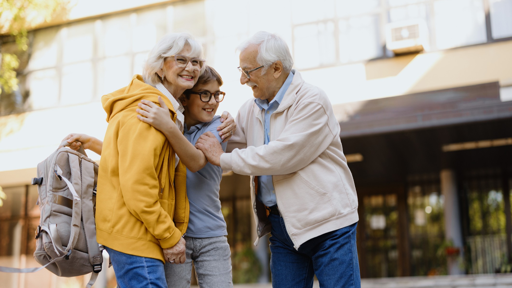 A boy with a backpack hugs his grandparents outside of school.
