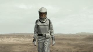 A woman wearing a protective silver suit walks in a wasteland