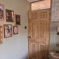 An oak door in a sage green bedroom with a variety of gold, framed prints on the wall.