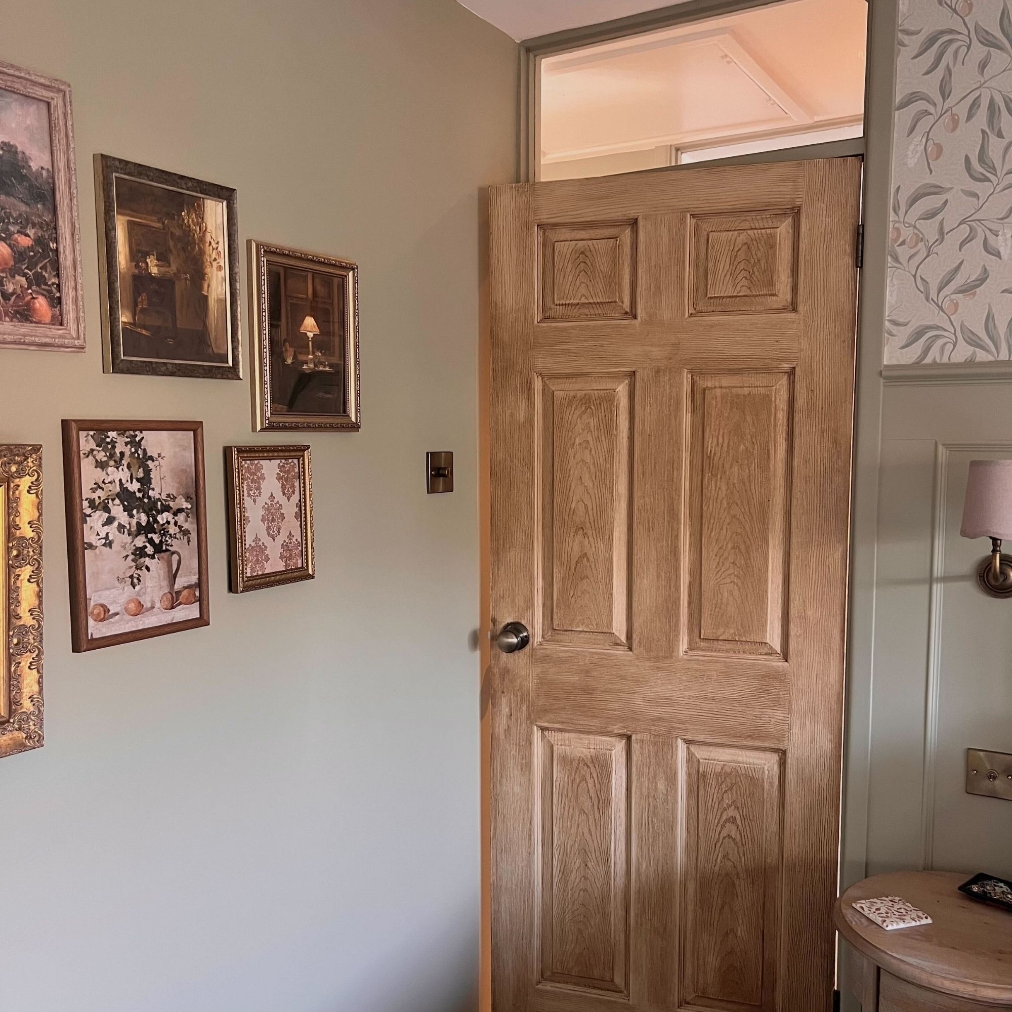 An oak door in a sage green bedroom with a variety of gold, framed prints on the wall. 