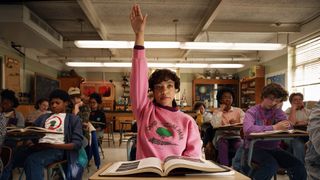 Priah Ferguson as Erica Sinclair in Stranger Things season 5 sitting in front of a school desk with an open book on it while raising her hand