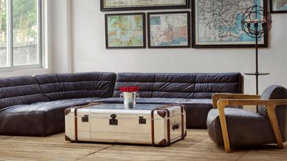 living room with leather curved sofa, timber floors, an armchair, gallery wall of maps, and metal storage trunk as the coffee table