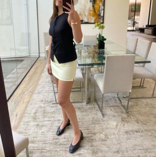 Woman wearing almond toe shoe trend with cream mini skirt and black top, taking mirror selfie.