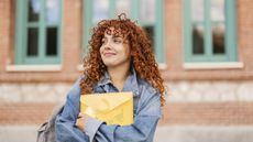 A college student smiles while she stands outside a building holding a manila envelope to her chest.