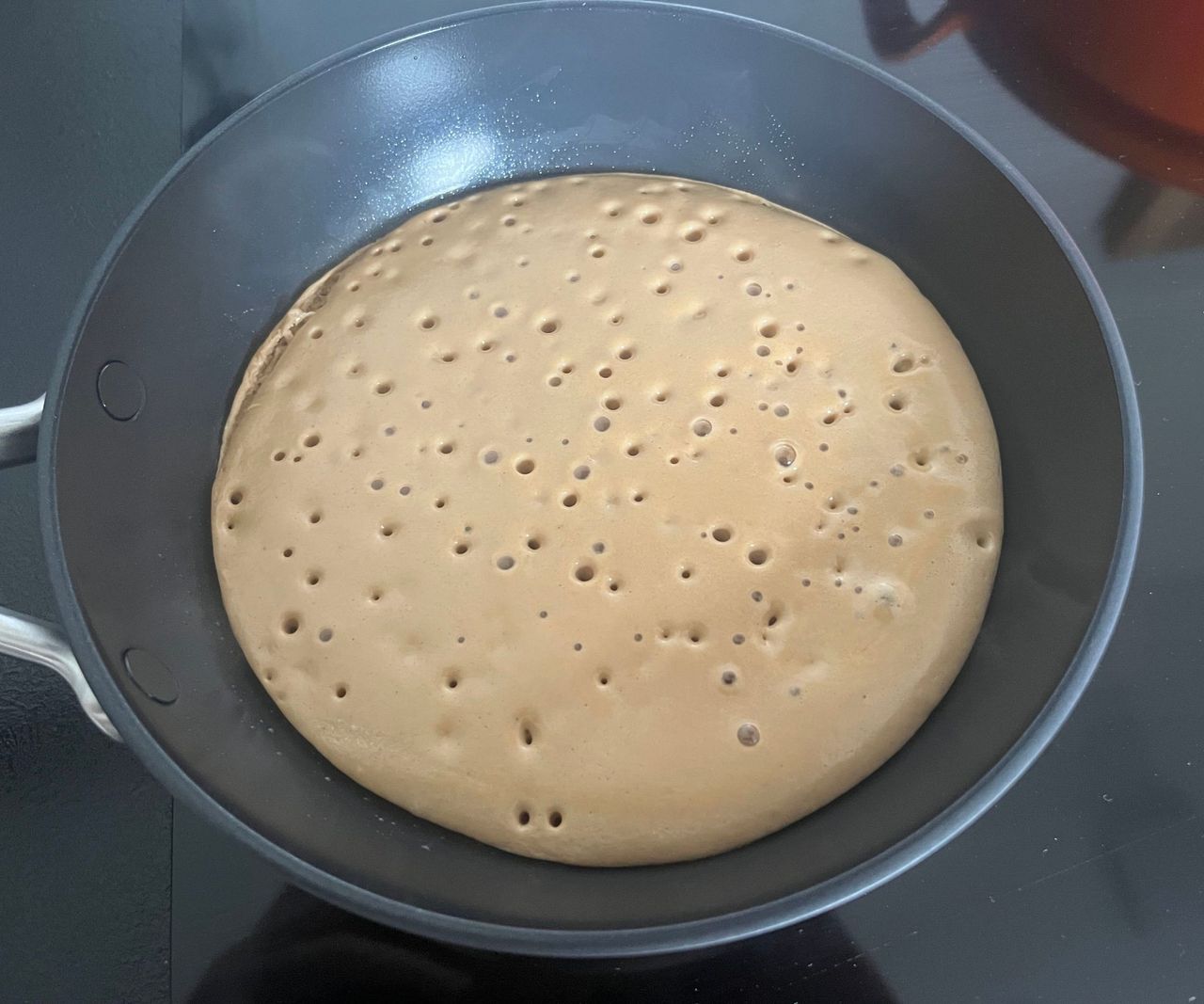 A black ceramic frying pan on a black stovetop with a pancake bubbling inside.
