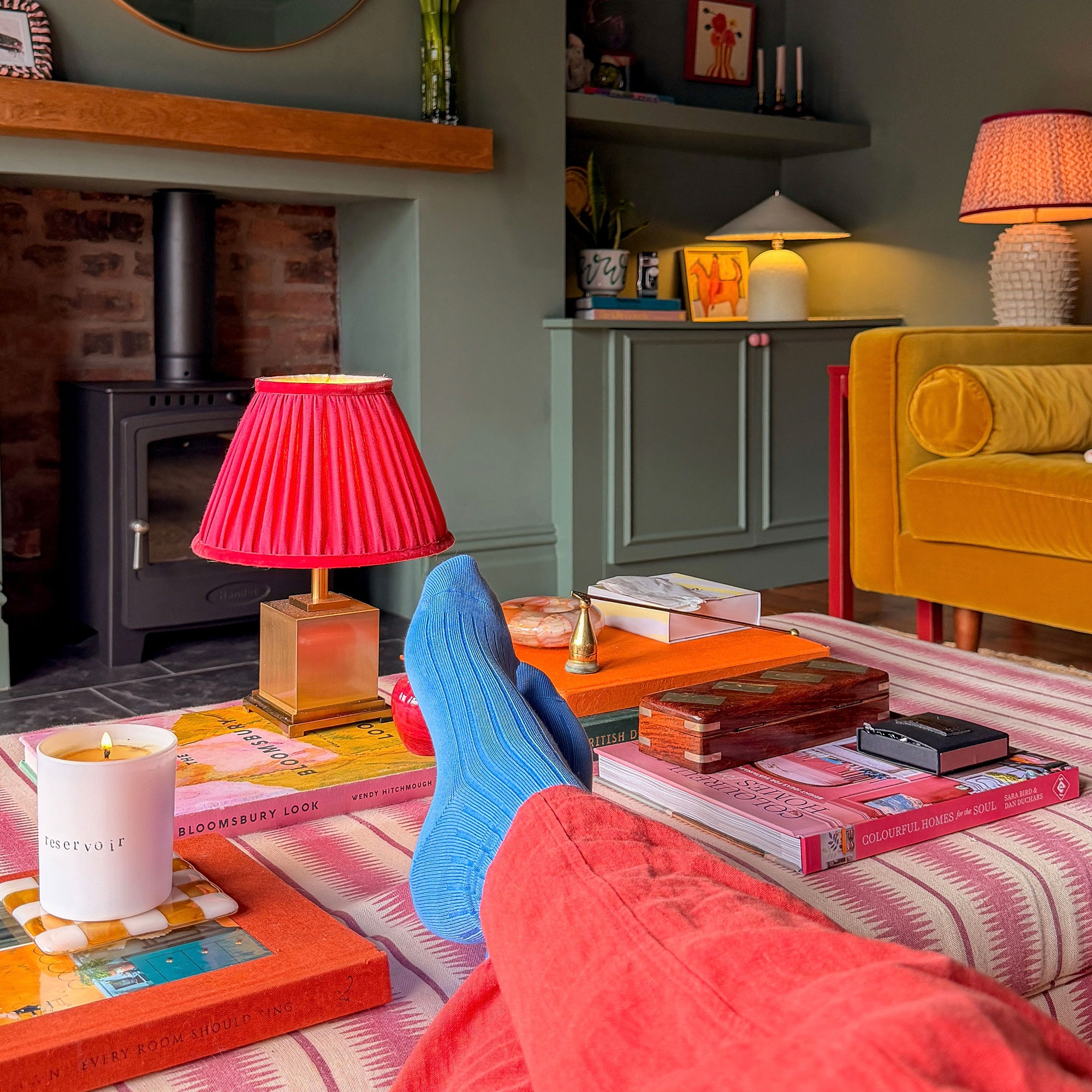 Woman in coral trousers and blue socks with feet resting on pink striped ottoman displaying candle, wireless lamp and stacks of coffee table books