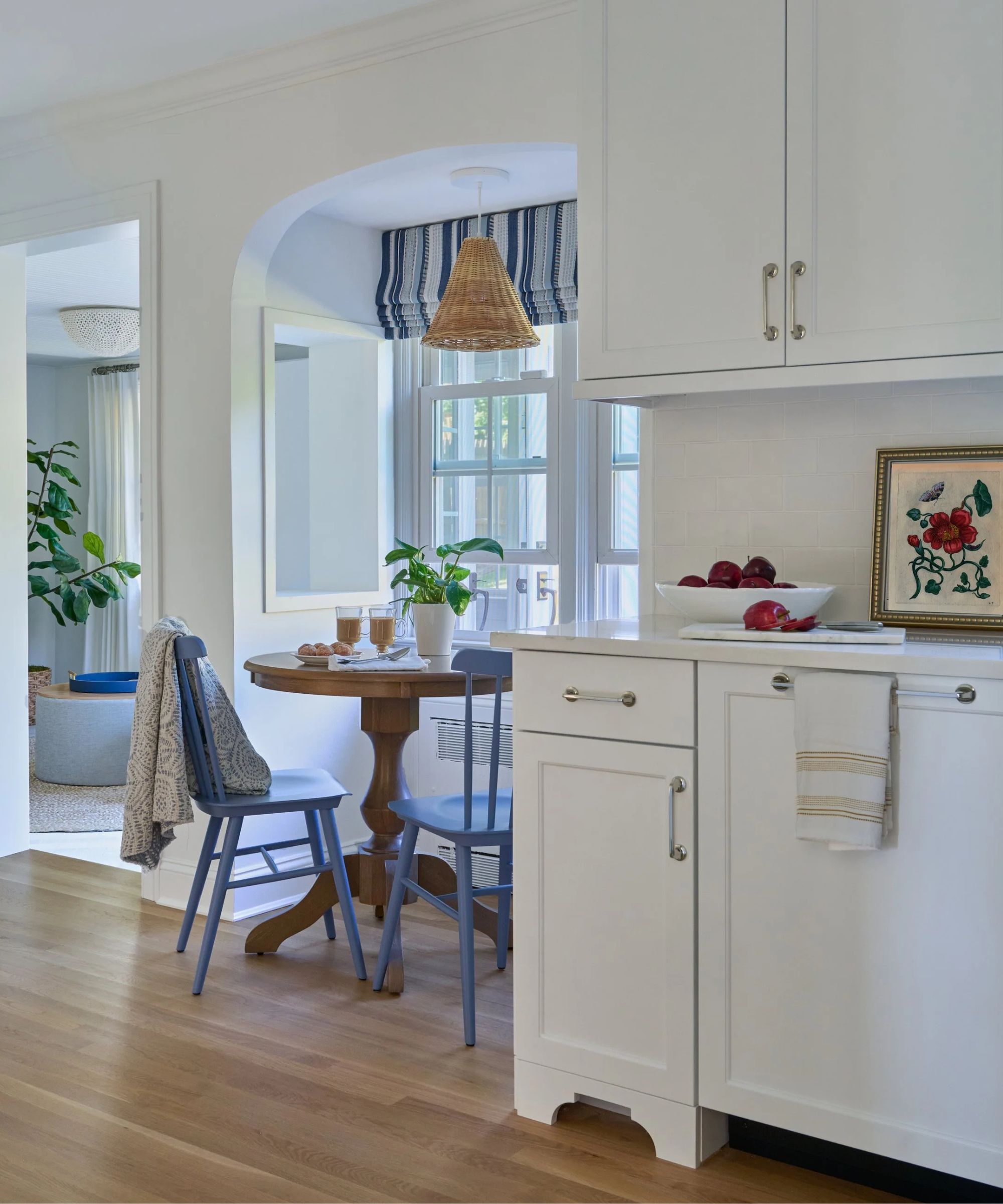 A white kitchen with an eat-in area added by the window, and dressed with blue striped window treatments, blue painted chairs, a wooden round table, a patterned throw, and a rattan pendant light