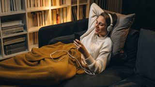 Woman listening to the best podcasts to fall asleep to on the sofa, wrapped up in a blanket with headphones on
