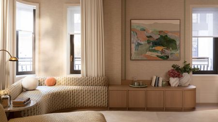 beige living room with curved yellow banquette seat under a window with white sheers, a large artwork over timber joinery styled with books, a plate of glasses, and two vases. on the sofa is two round cushions with a brass lamp