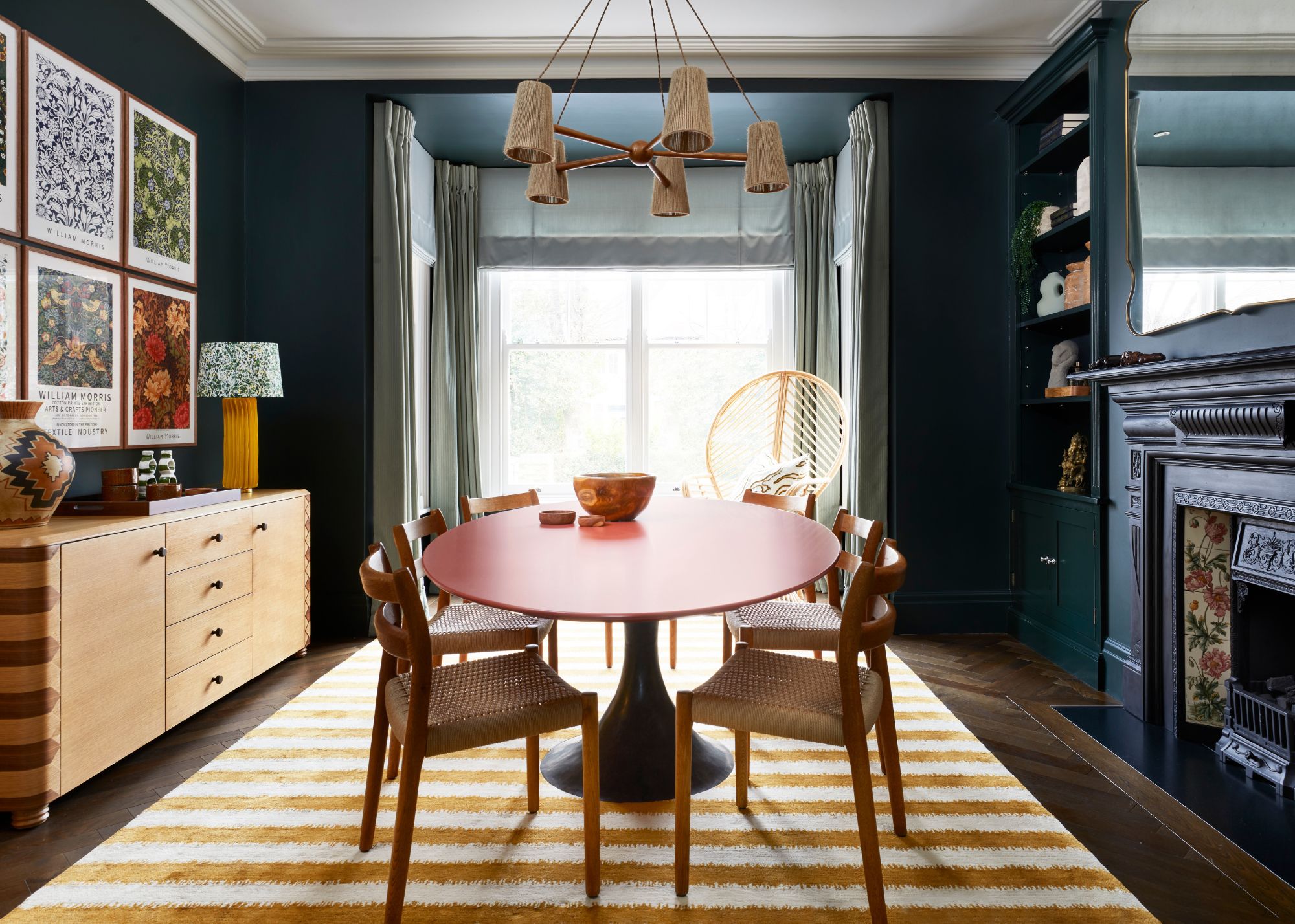 dining room with dark green walls, red dining table with bay window with light blue curtains and roman blind, yellow and cream striped rug
