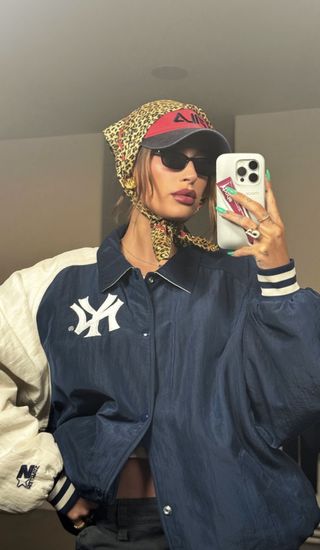 Hailey Bieber poses in front of a mirror with a leopard print scarf tied around her head in a Yankees jacket on the way to Coachella