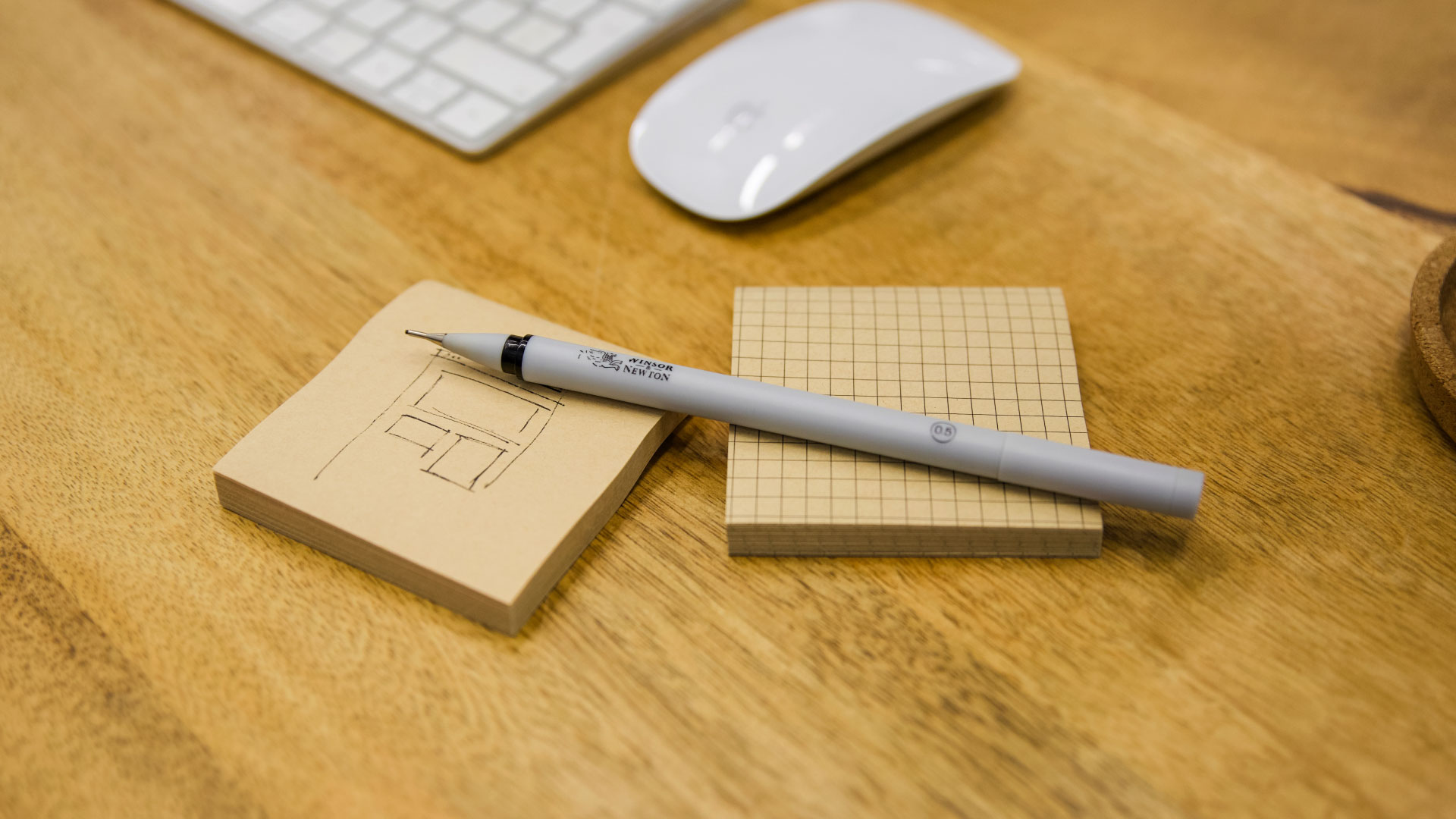 A gray drafting pen rests on two stacks of small, square, tan-colored, grid-patterned sticky notes, one of which has a simple architectural sketch.