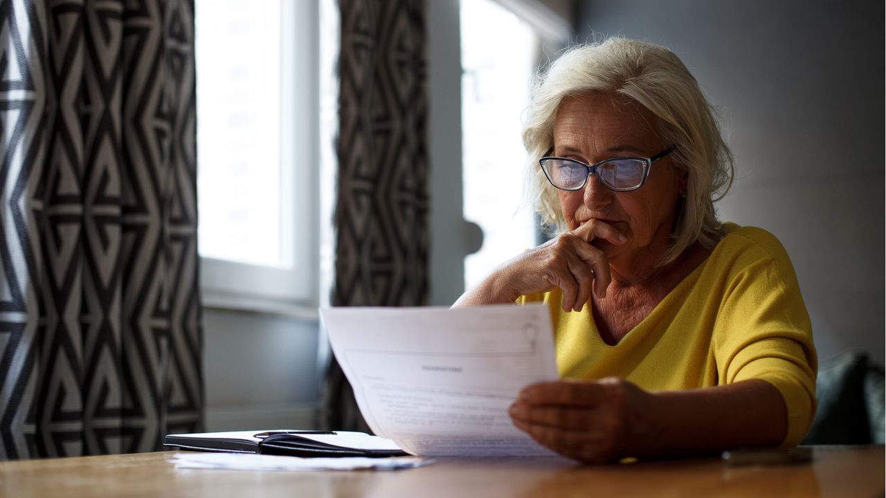 An older woman reads over paperwork while sitting at her dining room table.