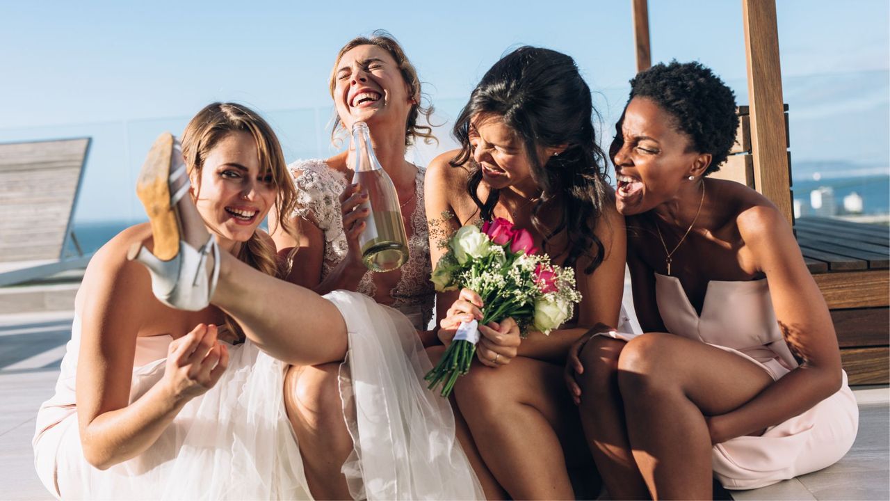 A bride and three bridesmaids sitting together on the beach laughing with flowers in their hand