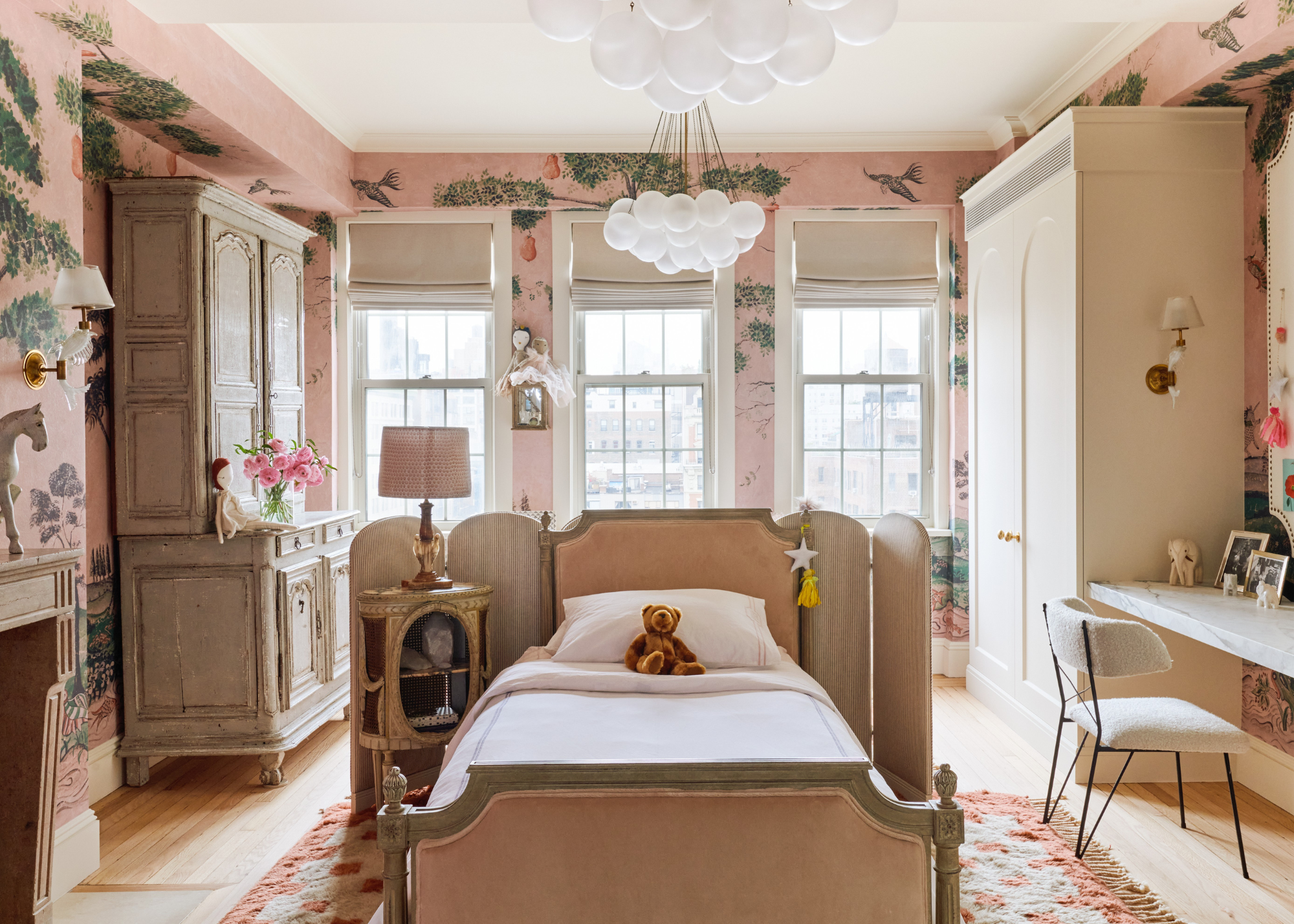 A kid's bedroom with a single bed in the center of the room, with a small room divider running along the back, and a small nightstand; pink patterned wallpaper covers all the walls, with one built-in and one standalone wardrobe unit, and a fitted desk with a boucle white chair