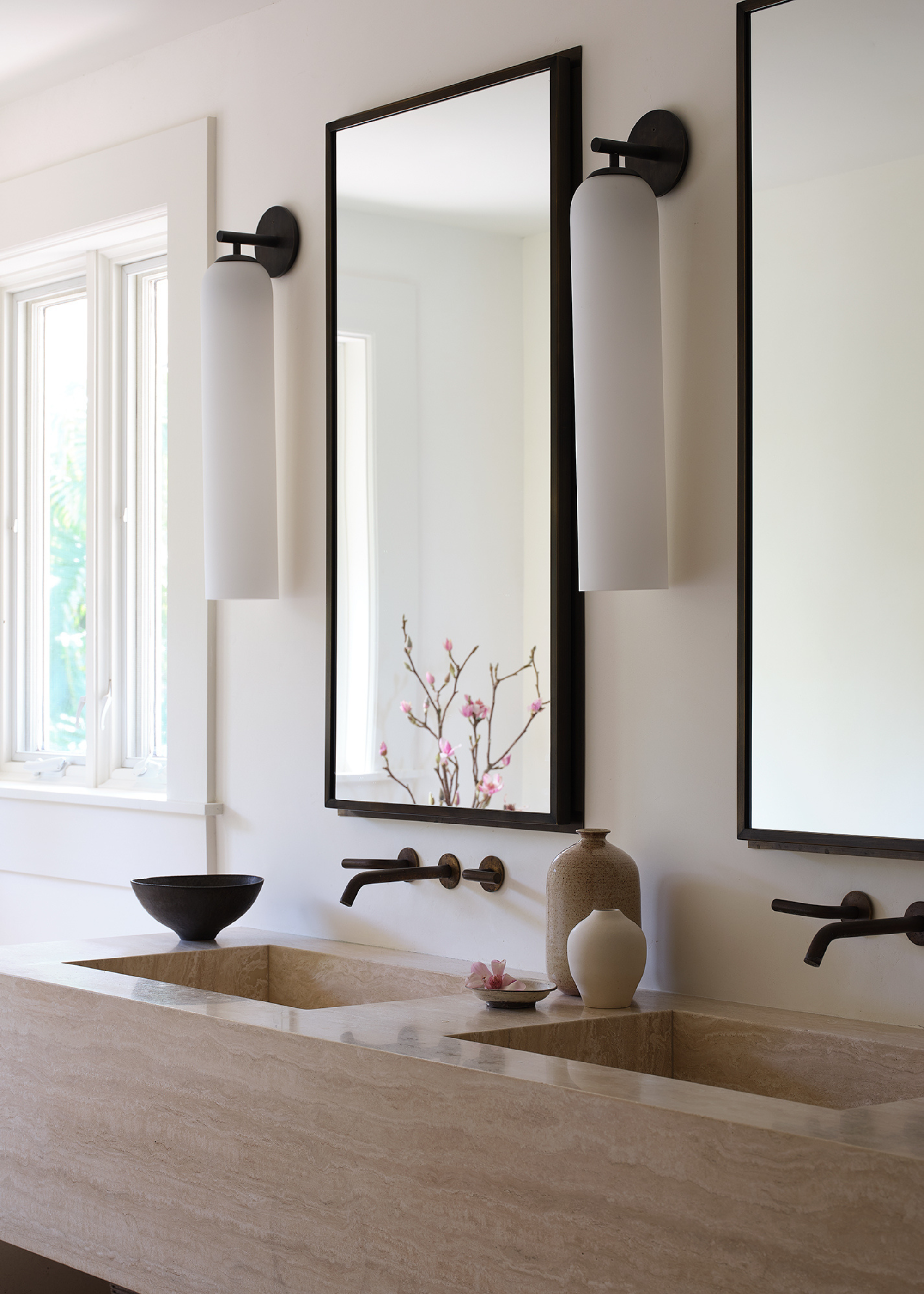 A calm bathroom with beige stone sinks, black hanging mirrors, vases, and wall lamps
