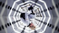 Male astronaut floating upside down in a tucked position in a space station. He has short blond hair and is wearing a gray jumpsuit.