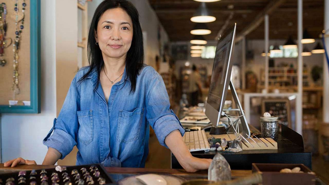 A woman stands at a workbench in her store, looking thoughtful.