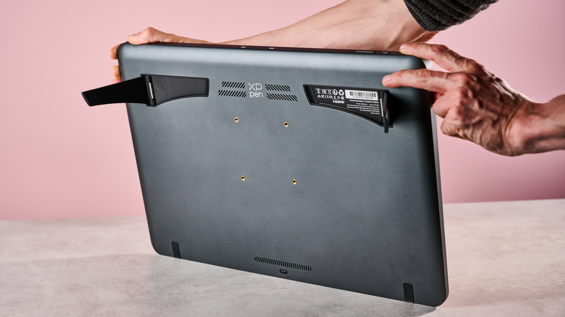 A man lifting up the XPPen Artist Pro 19 Gen 2 to show its feet in front of a pink background.